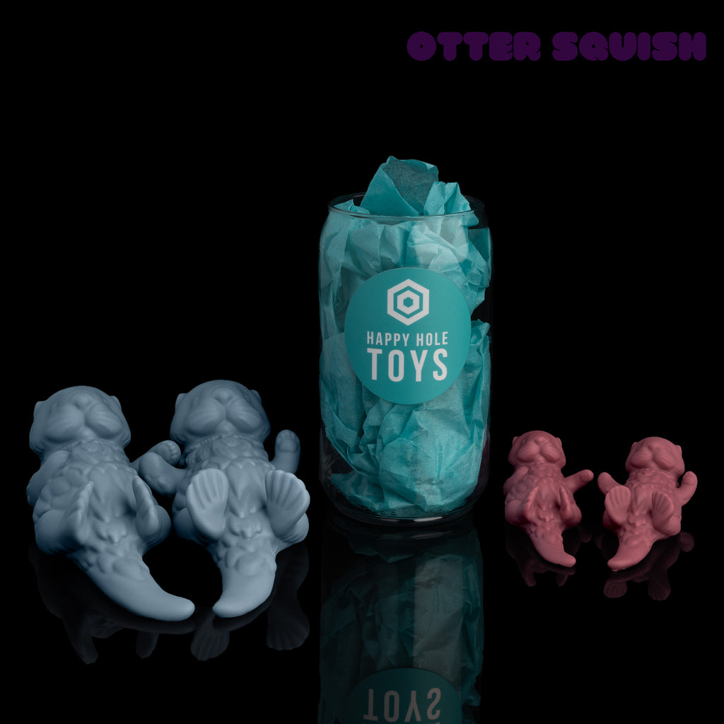 Two pairs of Otter Squishes with a clear plastic "can" filled with teal tissue paper in the middle for scale. On the left are the larger "buds" in Fjord, on the left are smaller "pups" in Raspberry.