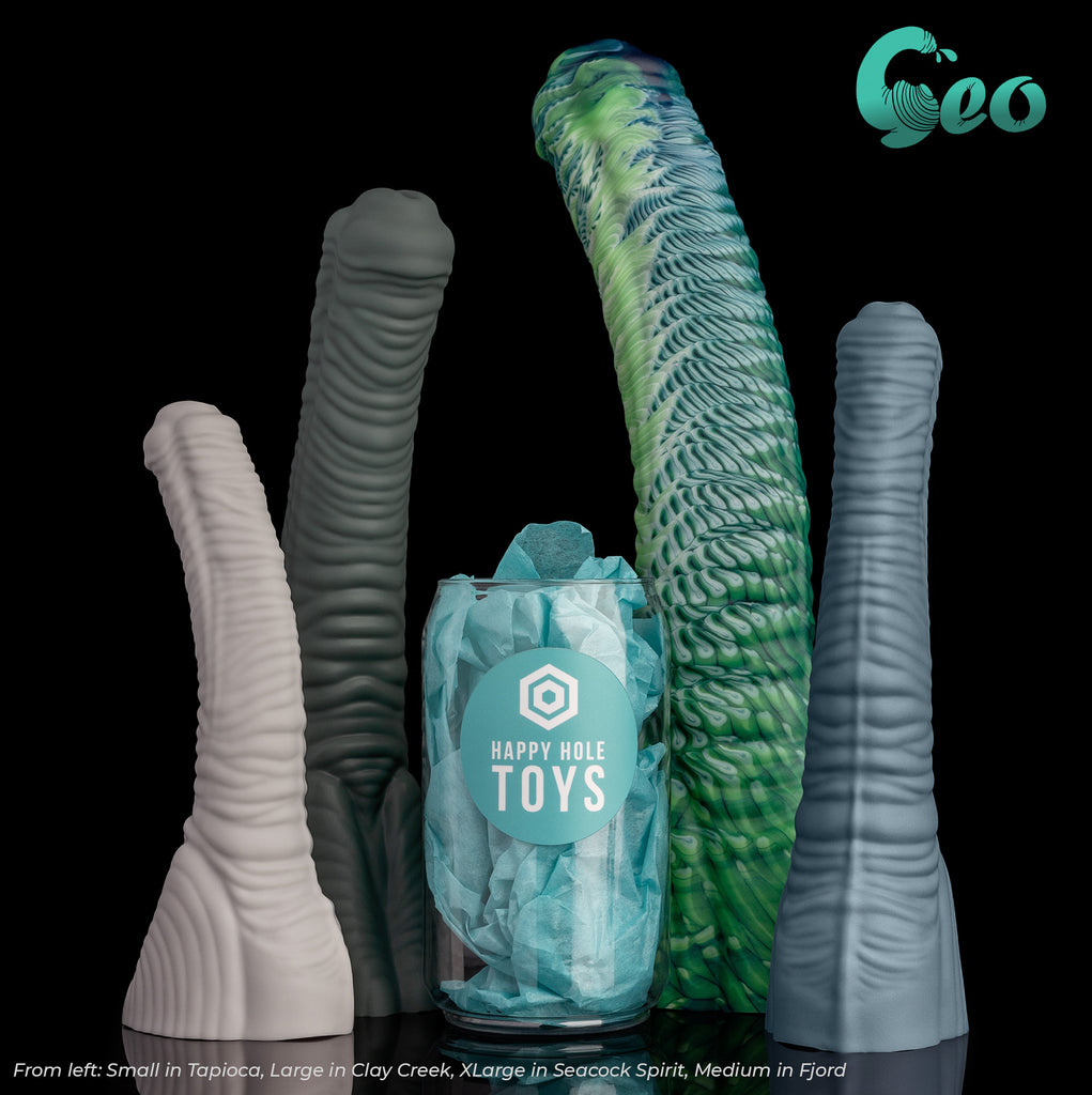 There are 4 Geo dildos in 4 different colors, three are in our Natural Series and one is a Premium Pour. There is a clear plastic "soda can" filled with teal tissue paper in the middle for scale. From left: Small in Tapioca, Large in Clay Creek, XLarge in Seacock Spirit (Premium Pour), and Medium in Fjord.