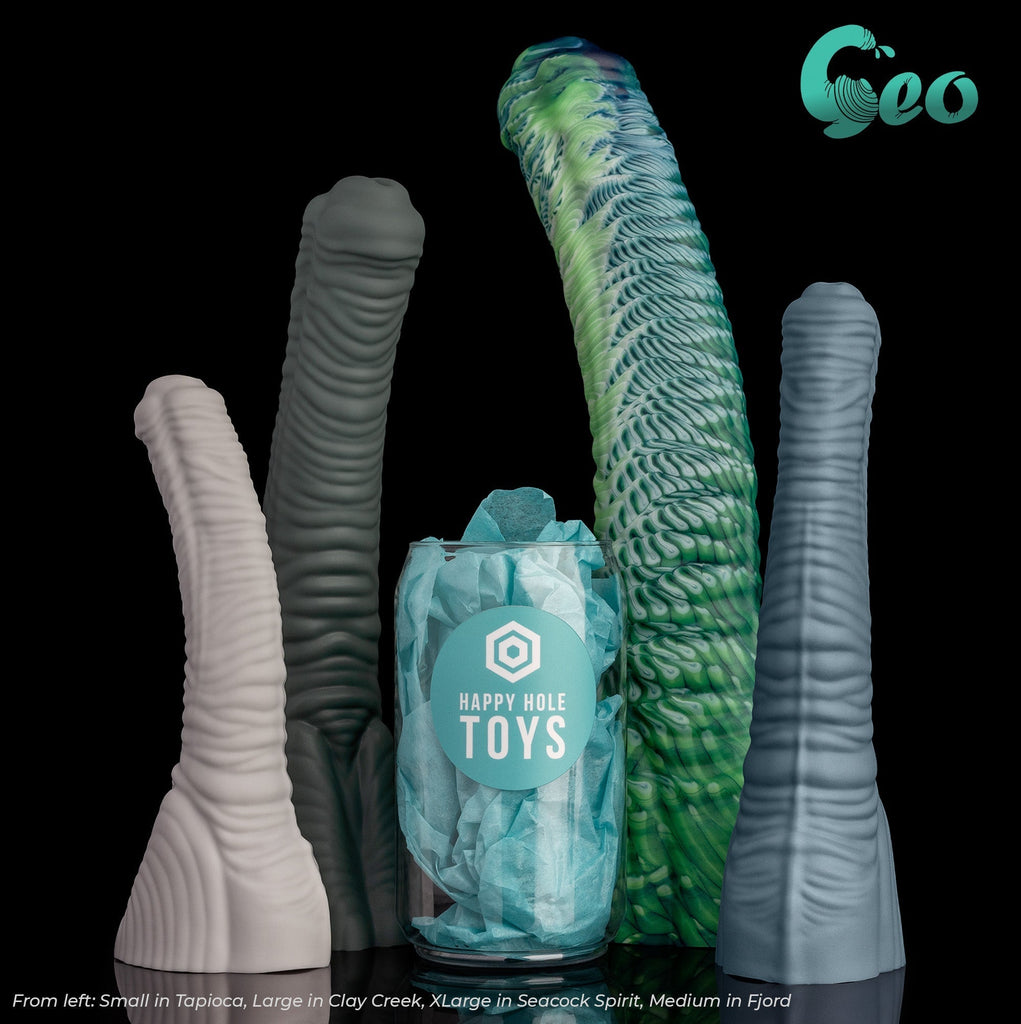 Geo size comparison: There are four Geo dildos in four different colors, three are in our Natural Series and one is a Premium Pour. There is a clear plastic "soda can" filled with teal tissue paper in the middle for scale. From left: Small in Tapioca, Large in Clay Creek, XLarge in Seacock Spirit (Premium Pour), and Medium in Fjord.